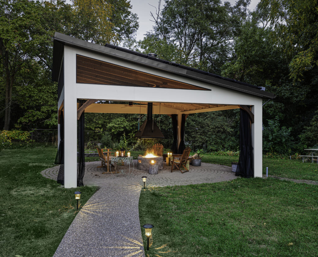 A captivating image showcasing a stand-alone covered pavilion, its architectural elegance sheltering a cozy seating arrangement. A central firepit emanates a gentle, comforting glow, surrounded by an aura of open space and natural beauty.